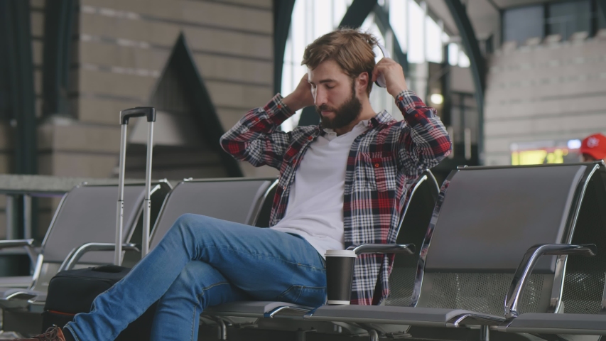 Hipster traveler relaxing listening to music in headphones while waiting for his train in waiting hall. Youg bearded passenger sitting in airport terminal waiting for flight putting on earphones
