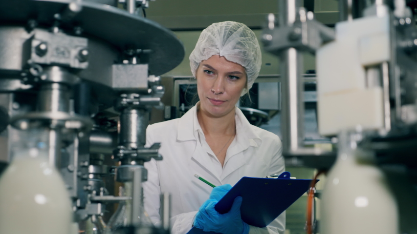 A woman checks bottles with milk, moving on a conveyor. Quality control of production line at a factory. Female worker controls the quality of production equipment. Royalty-Free Stock Footage #1042712404
