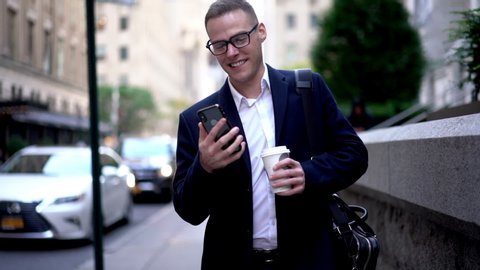 Formally dressed businessman walking with coffee to go and smartphone device for browsing in slow motion, successful male trader drinking hot caffeine beverage while walking at city street
