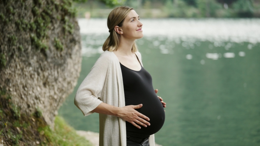 Pregnant woman touching gently her tummy when standing near the mountain lake.
