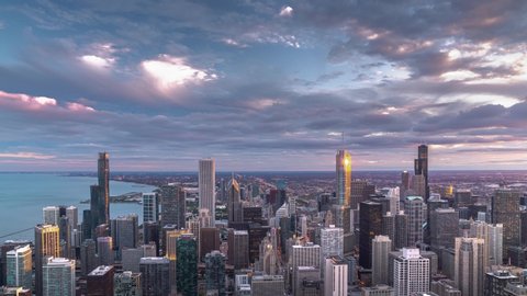 A beautiful aerial skyline panoramic sunset day to night zoom in time lapse of downtown Chicago with Lake Michigan and colorful pink and blue clouds as the sun sets and lights come on in the city.