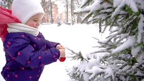 Cute little baby girl hanging red Christmas ball on snowy branches of green fir tree outdoors growing in city park. Slow motion full hd video footage.
