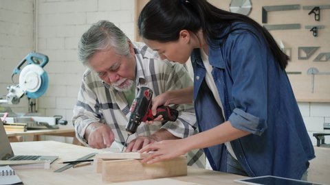 Asian senior male and teenage girl carpenter building modern furniture in home workshop. Man and woman use electric drill to drill wood. Concept of work business from home