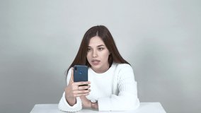 Light gray background Caucasian girl blogger white jumper Straight long hair sitting at a table, holding a phone, saying something to the camera, angry, annoyed