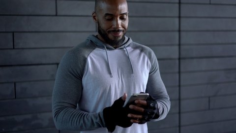 Young african american sportsman in active wear using smartphone app for checking fitness activities and workout standing on wall background
