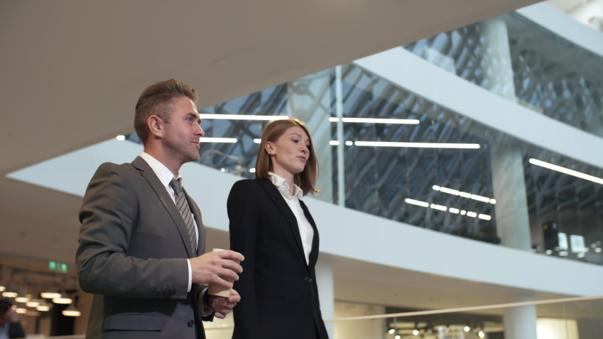 Side view follow medium shot of two Caucasian businesspeople, man and woman, walking through office building and talking