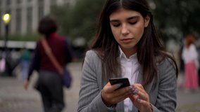 Serious attractive spanish millennial female using cellular for sending text messages and chatting in social networks standing outdoors
