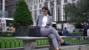 Confident spanish businesswoman millenial in formal wear and trendy eyewear using mobile phone sitting on bench in metropolis with bag
