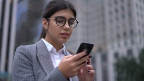 Positive spanish millennial female manager in glasses chatting in social networks via messengers apps on smartphone outdoors using 4G connection
