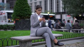 Happy successful millennial female in formal wear and glasses checking message on smartphone via application spending time outdoors
