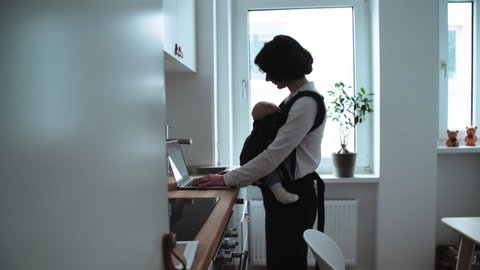 Woman standing in kitchen using laptop and with baby sleeping in carrier.