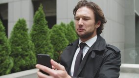 Handsome young businessman with fair hair and beard wearing a dark coat and tie talking on smartphone. He is unhappy and argues with the interlocutor.