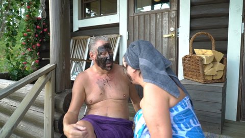 The elderly couple are happy together sitting on the wooden steps of the Finnish sauna and using black facial cosmetic peeling masks. They acting all silly and happy laughing