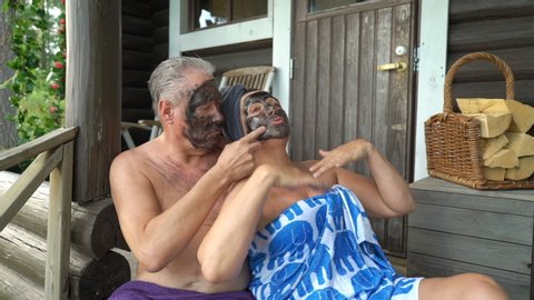 The elderly couple are happy together sitting on the wooden steps of the Finnish sauna and using black facial cosmetic peeling masks. They acting all silly and happy laughing