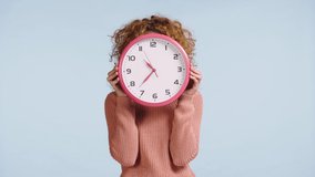 cheerful woman looking out from behind clock isolated on grey