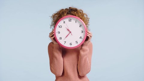 cheerful woman looking out from behind clock isolated on grey