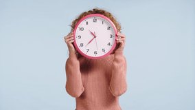 displeased woman hiding face behind clock isolated on blue