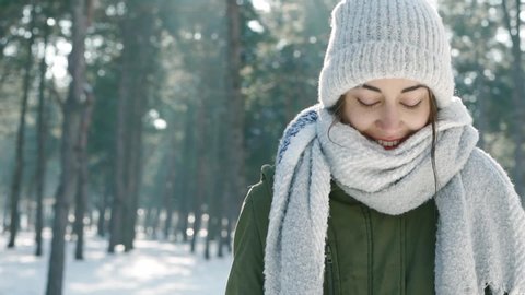 slow motion closeup portrait of beautiful laughing young woman in warm woolen cap and long scarf in snowy winter park at frozzy sunny day. woman looks playful, warming hands and wraps herself 