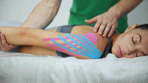 Doctor helps woman that lying down by moving her hand with kinesio tape on it.