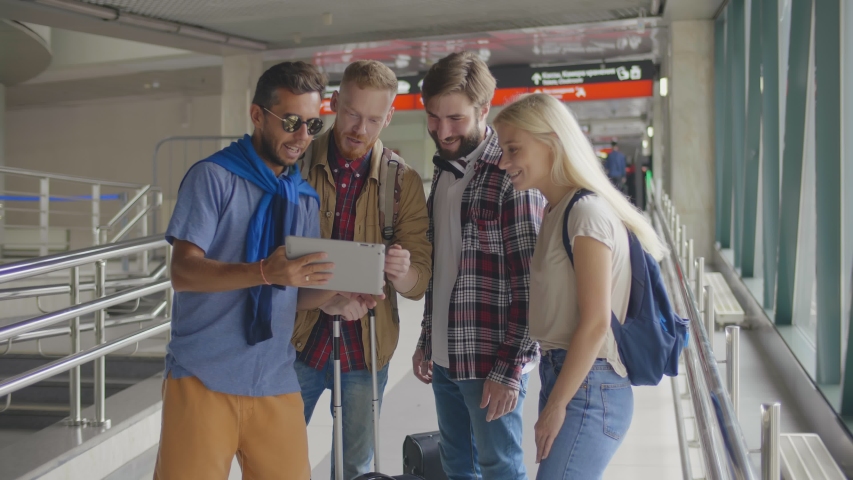 Young friends gathered together and reading information about their trip on digital tablet standing in airport terminal. Youg people choosing booking tickets on tablet