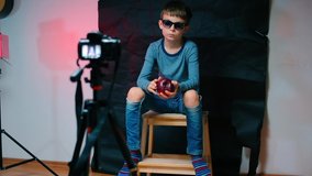 An independent child makes a video blog. Holds an apple in his hands, bites it. There are ripped jeans on his feet. He waves his hand at the camera.