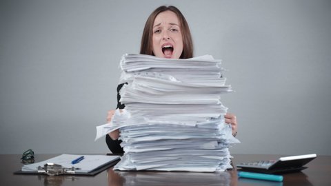 Stressed female office worker sitting at the table behind the pile of paperwork and looking withsad face on camera. Woman begging for help, stack of paper files, unfilled documents and application