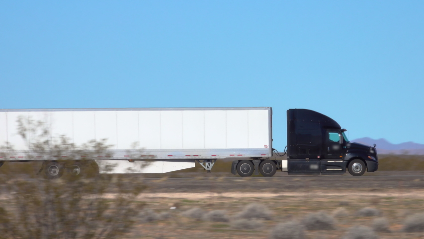 CLOSE UP: Black semi-trailer truck speeds along the interstate highway crossing a desert in United States. Cargo lorry hauls heavy freight container across the countryside of United States of America. Royalty-Free Stock Footage #1042796074