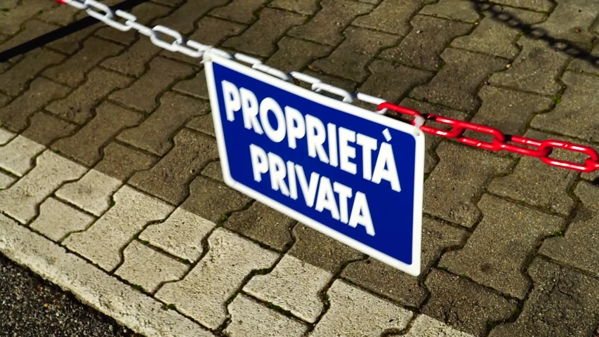 red and white chain with signal with "private property" written in Italian on a blue background and geometric pavement with shadow