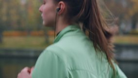 Side view of beautiful sporty girl in earphones thoughtfully running in autumn city park on riverside