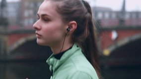 Portrait of beautiful sporty girl in earphones confidently running in autumn city park on riverside