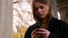 Attractive serious girl in coat thoughtfully using cellphone walking  outdoor at autumn