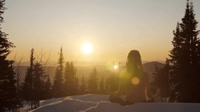 Slow mo video of  Young female wearing long hair dreads, seating in yoga lotus pose throwing snow and meditating on sunrise in snow forest on mountain top. Cold frozen winter morning