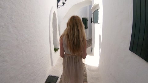 Blonde unrecognizable woman on her trip walking by the beautiful southern street in summer. Beautiful slow motion shot in Menorca most  famous   fisherman village Binibeca. White old houses.