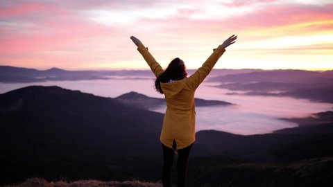 Young beautiful woman stands on top of a mountain and raises her hands up to meet the sunset. Hiker woman standing with hands up achieving the top and enjoys incredible mountain landscape