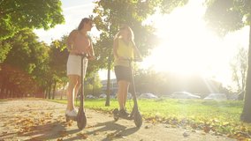 SLOW MOTION, LOW ANGLE, LENS FLARE: Golden autumn sun rays shine on two female friends riding electric scooters around the green park. Cheerful women cruise down the avenue on new electric scooters. - Powered by Shutterstock - Get 15% off with code: PIKWIZARD15