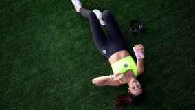 Top view of young female runner lying on green grass for recreating and taking selfie images for sending media message during mobile communication, muscular athlete making photo outdoors
