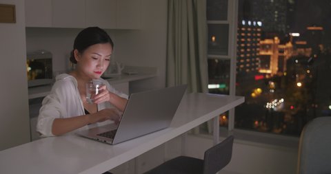 asian young serous businesswoman siting by table working at night on computer typing and thinking carefully looking at laptop holding water cup at home or modern hotel 스톡 비디오