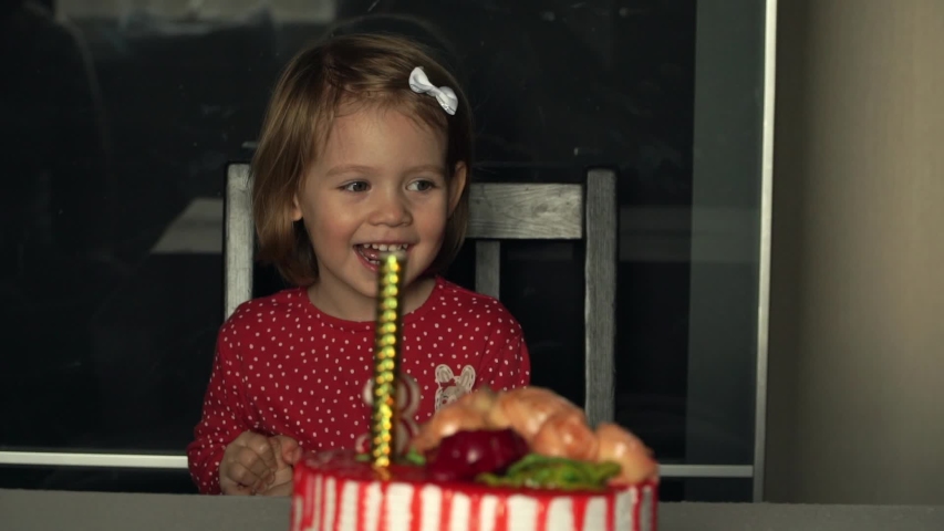 Close up of portrait of little smiling child girl celebrates birthday with festive cake with cream, candles, laughs and looks with delight how to set fireworks on fire. Happy holiday concept