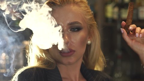 Close-up of a woman in a business suit Smoking a cigar in a bar, in the background a bar counter with bottles of alcohol. Lots of smoke from a cigar or cigarette. Nightlife. Slow motion.