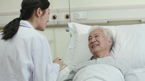friendly young asian female doctor talking to male senior patient at bedside in hospital ward