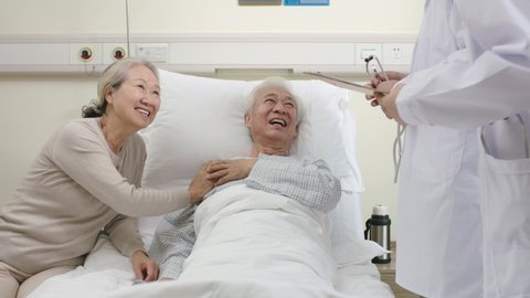 two young asian doctors making rounds talking to senior patient in hospital ward