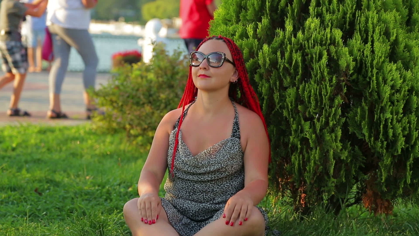 Young tanned woman in sunglasses meditates under a tree in a park