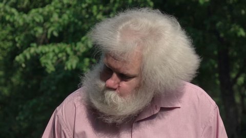 Bearded senior man with splendid long hair shaking his head on green tree background. Close up funny outdoor portrait of handsome person in slow motion at sunny day. Santa Claus in summer holiday.