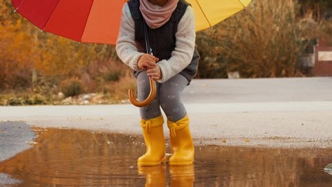 Happy Little Girl with A Multicolored Umbrella Jumping On Puddles In Yellow Rubber Boots. Slow Motion