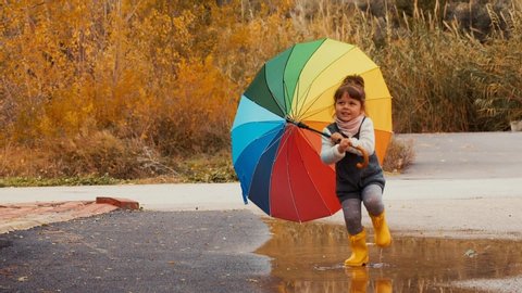 Happy Little Girl with A Multicolored Umbrella Jumping On Puddles In Rubber Boots. Slow Motion