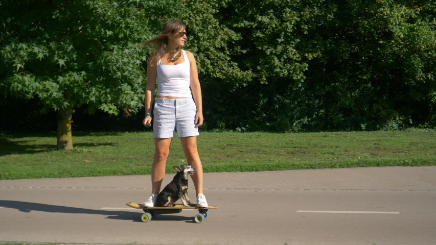 Modern girl rides an e-longboard through the scenic green park with her miniature pinscher dog. Joyful Caucasian woman enjoys a fun cruise on e-skateboard with her cute little puppy. Royalty-Free Stock Footage #1042889938