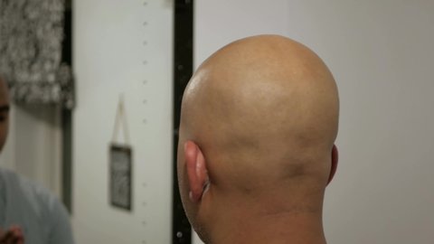 Closeup of a bald headed brown asian man dabbing post shaving oil to a clean shaven head in front of a mirror