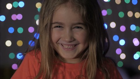 Cute Little Girl With Standing Against Glowing Christmas Lights Smiling at Camera. Portrait Child Looking at Camera. Christmas New Year Background. Lovely Girl Posing On Christmas Lights Background