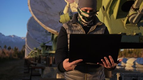 Hacker hacks the radio telescope using notebook. Man to program hacking on laptop in nature. Terrorist is hiding in camouflage balaclava. Hacker has criminal cases on the Internet. Programmer uses a