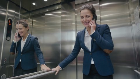 Attractive brunette business lady going up in modern steel elevator in large office center and talking on cell phone. Young businesswoman talks by smartphone and leaves elevator